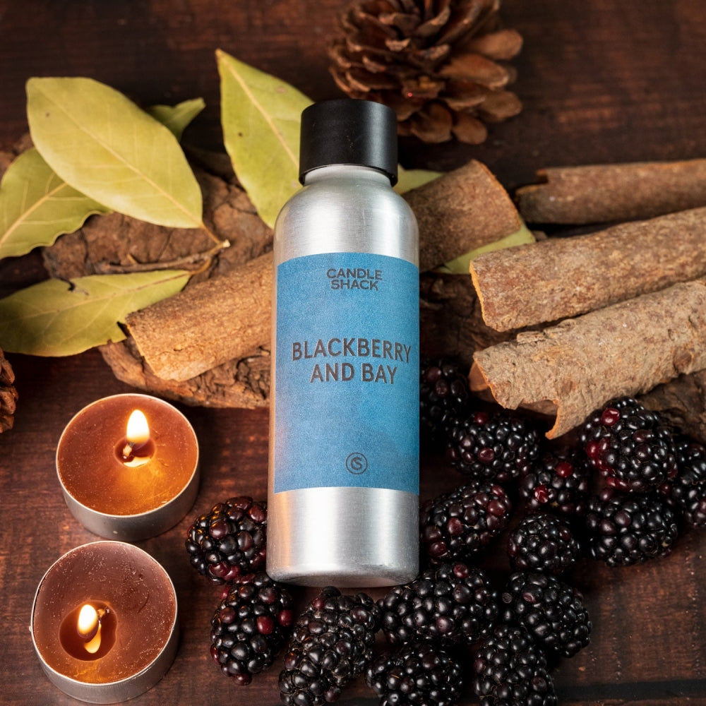 Sølvfarget flaske med blå etikett "Blackberry and Bay" omgitt av bjørnebær, kanelstenger, laurbærblader og telys