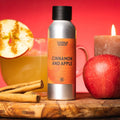 Sølvfarget flaske med oransje etikett "Cinnamon and Apple" ved siden av epler, kanelstenger og et tent lys.