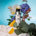 Sølvfarget flaske med Clean Cotton duftolje, omgitt av appelsinskiver, grønne blader og blomster på hvit duk.