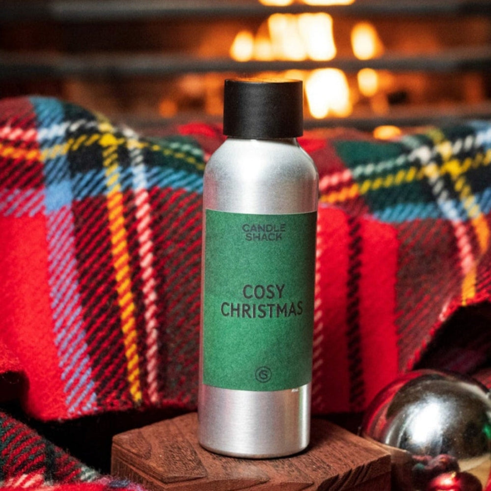 Sølvfarget flaske med grønn etikett for Cosy Christmas duftolje, foran et rutete pledd og peis.