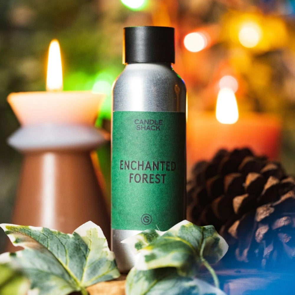 Sølvfarget flaske med grønn etikett "Enchanted Forest" duftolje, foran et tent lys og kongle