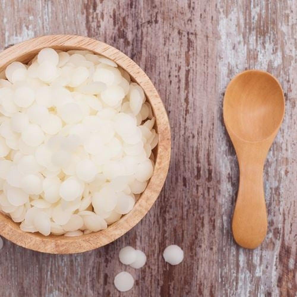 Hvit bivoks i pastiller i en trebol, med en treskje og noen løse pastiller på et treunderlag