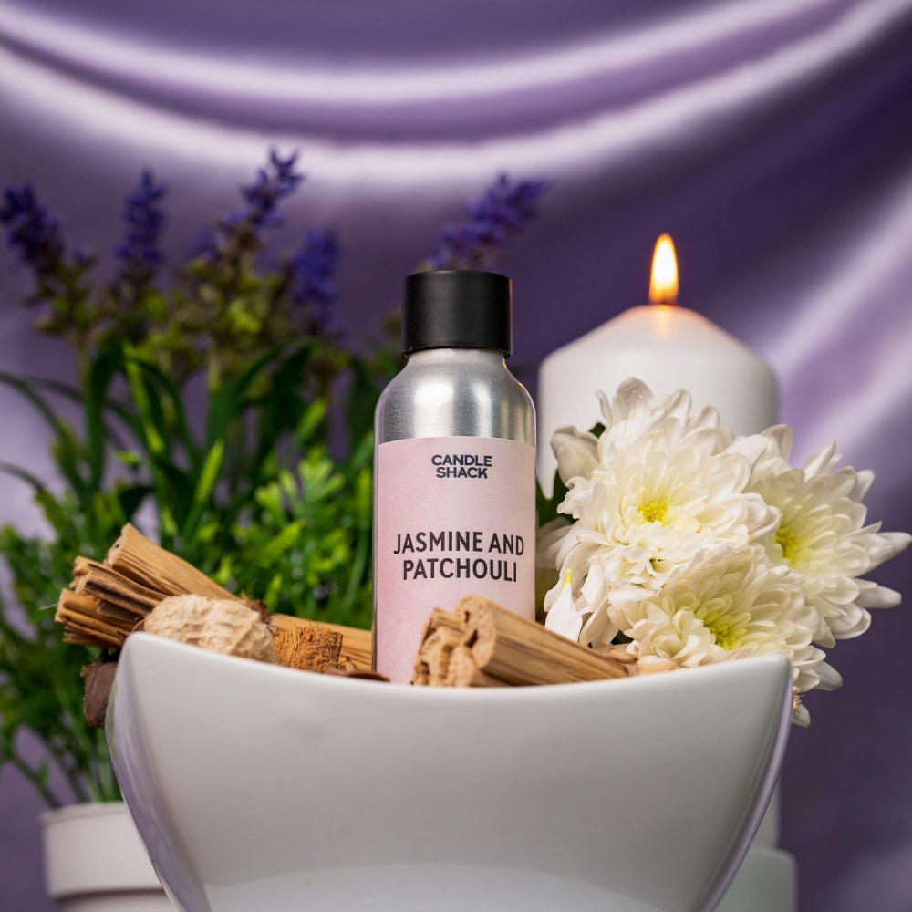 Sølvfarget flaske med Jasmine & Patchouli duftolje, omgitt av hvite blomster, kanelstenger og et brennende lys
