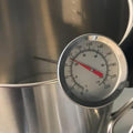 Sølvfarget klokketermometer med rød viser, festet til en metallkjele, viser temperatur i Celsius og Fahrenheit.