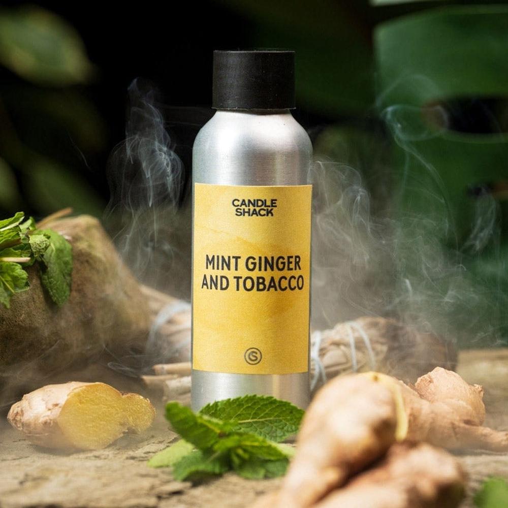 Sølvfarget flaske med svart kork og gul etikett for Mint, Ginger and Tobacco duftolje, omgitt av mynte og ingefær
