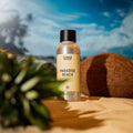 Paradise Beach duftolje i sølvflaske med svart kork, på sand med kokosnøtt og ananasblad, mot blå himmel