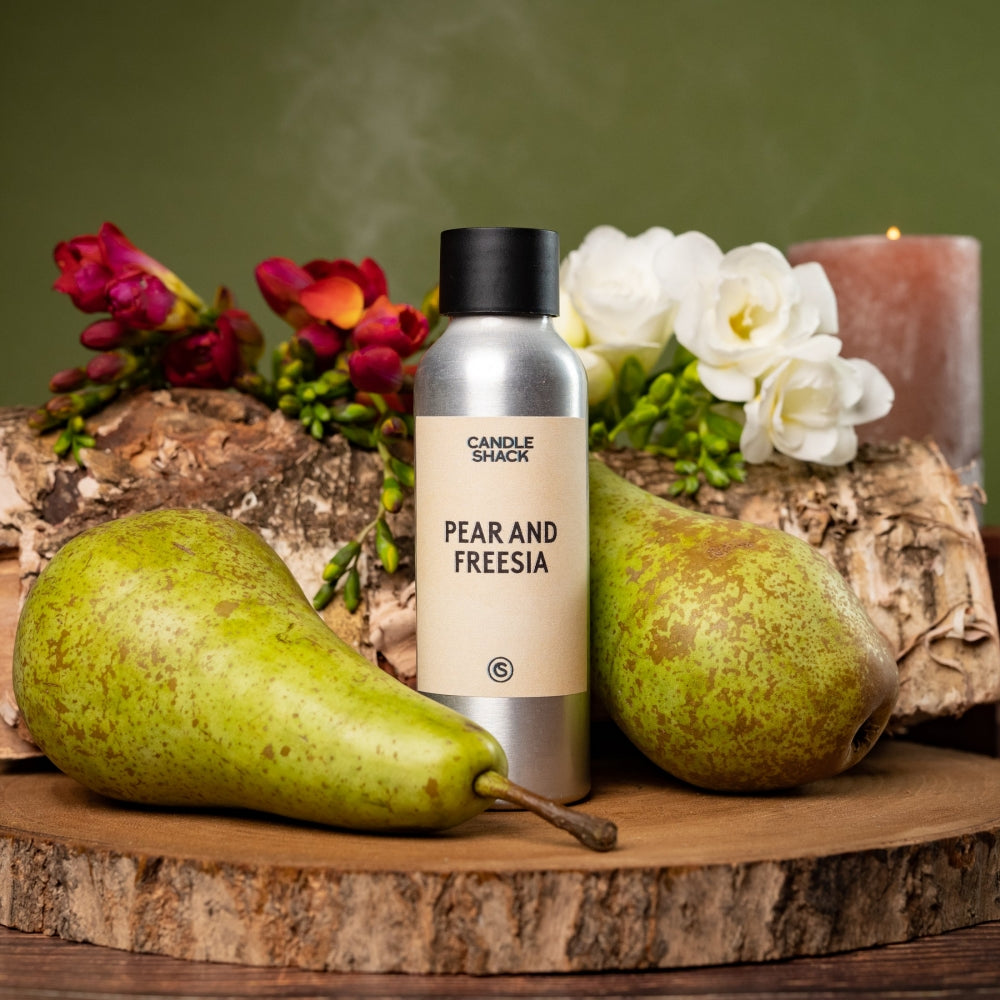 Sølvfarget flaske med Pear & Freesia duftolje, omgitt av grønne pærer, fresia og bjørkestokker på en treplate