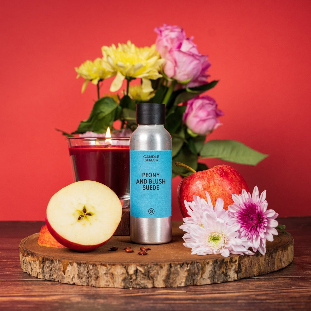 Sølvfarget flaske med Peony and Blush Suede duftolje, rødt lys, epler og blomster på trestubbe