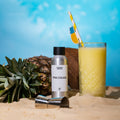 Pina Colada duftolje i sølvflaske, ananas, kokosnøtt og cocktailglass på sand med blå bakgrunn
