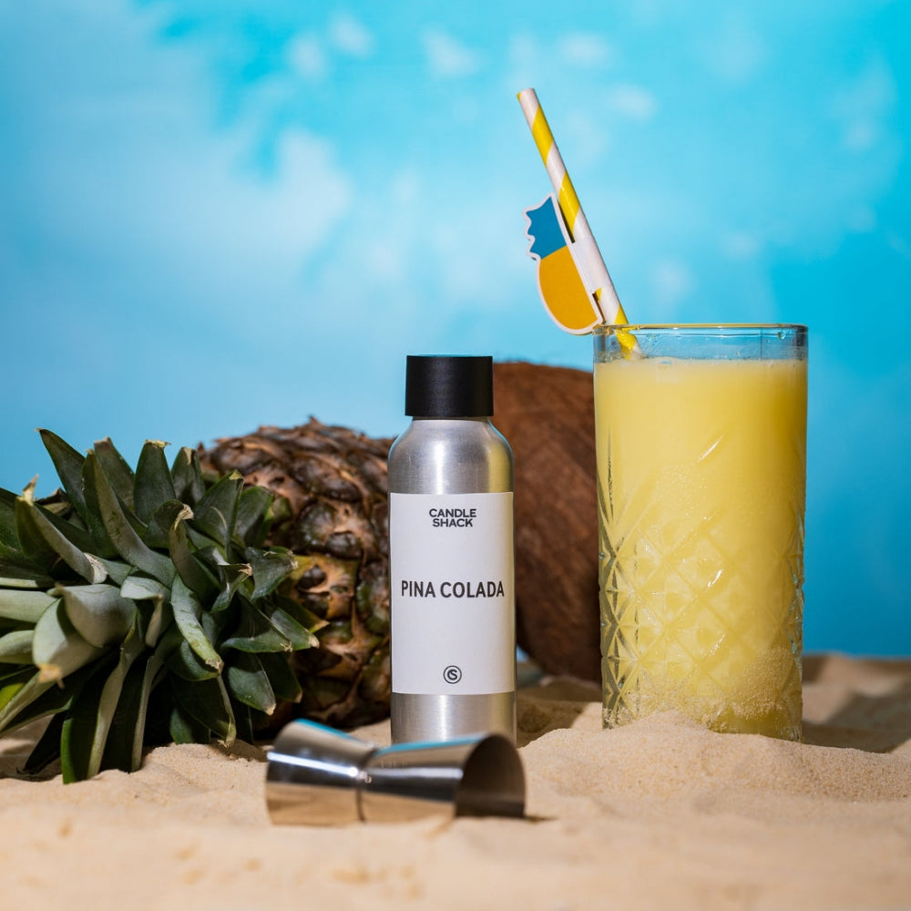 Pina Colada duftolje i sølvflaske, ananas, kokosnøtt og cocktailglass på sand med blå bakgrunn