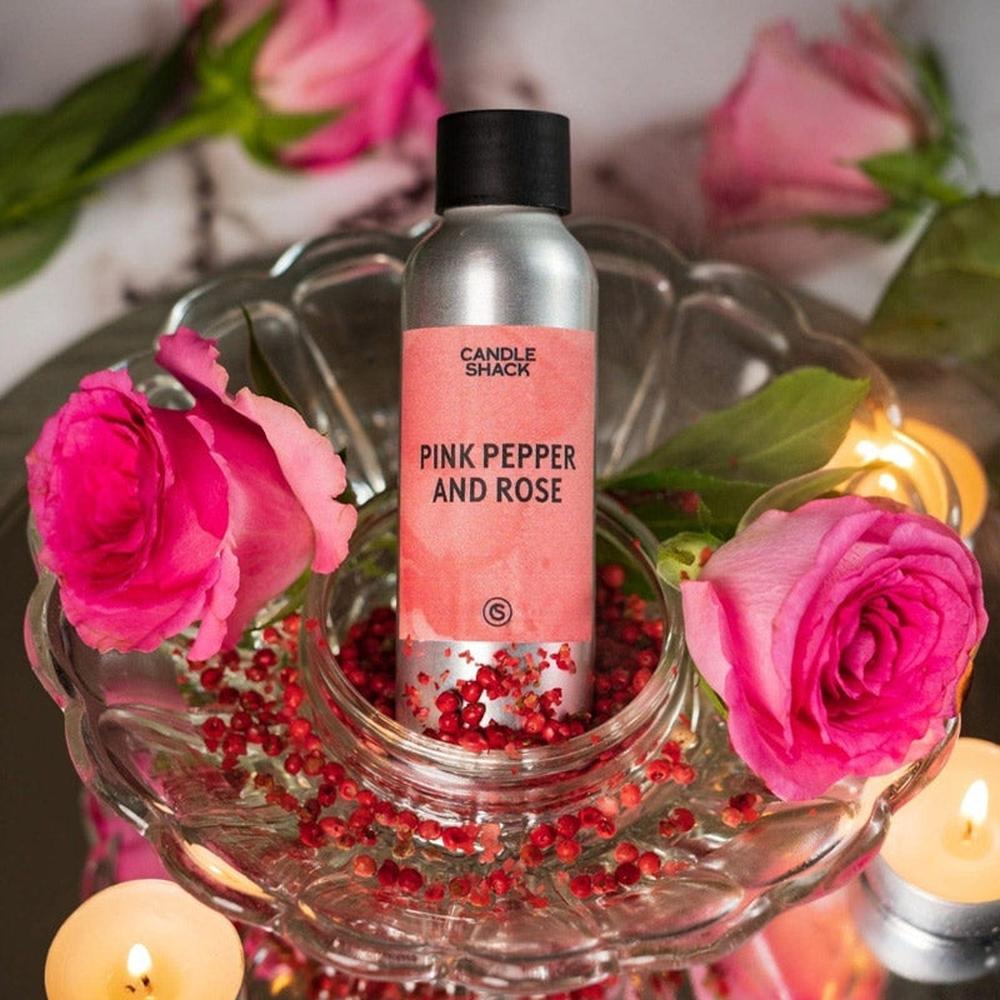 Sølvfarget flaske med Pink Pepper and Rose duftolje, omgitt av rosa roser, røde pepperkorn og tente telys