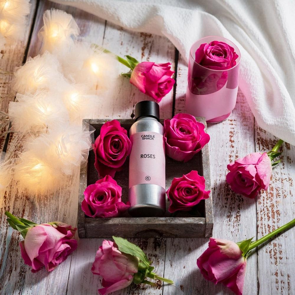 Sølvfarget duftoljeflaske "Roses" med rosa etikett, omgitt av friske rosa roser på trebrett og hvit duk