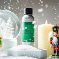 Sølvfarget flaske med grønn etikett "White Christmas" duftolje, omgitt av snø, et tent lys og julepynt.