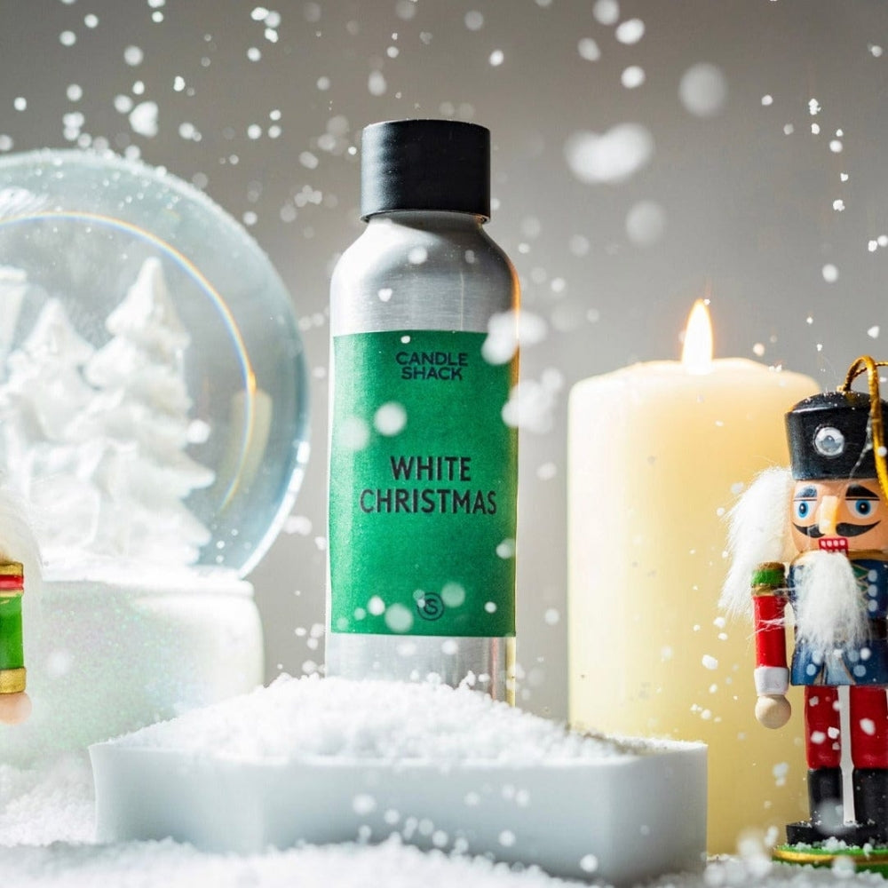 Sølvfarget flaske med grønn etikett "White Christmas" duftolje, omgitt av snø, et tent lys og julepynt.